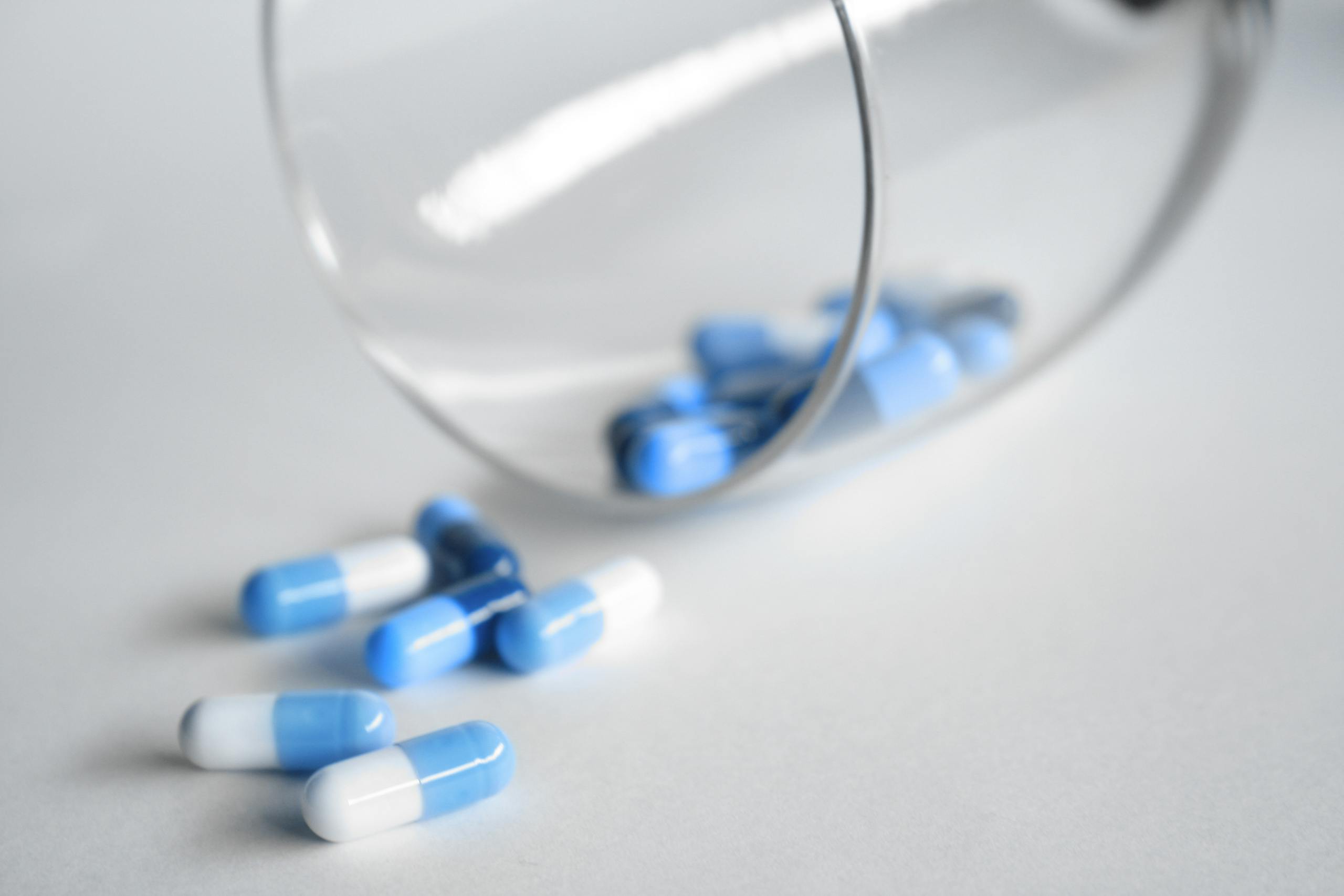 A close-up of blue and white capsules spilling from a glass on a light background.